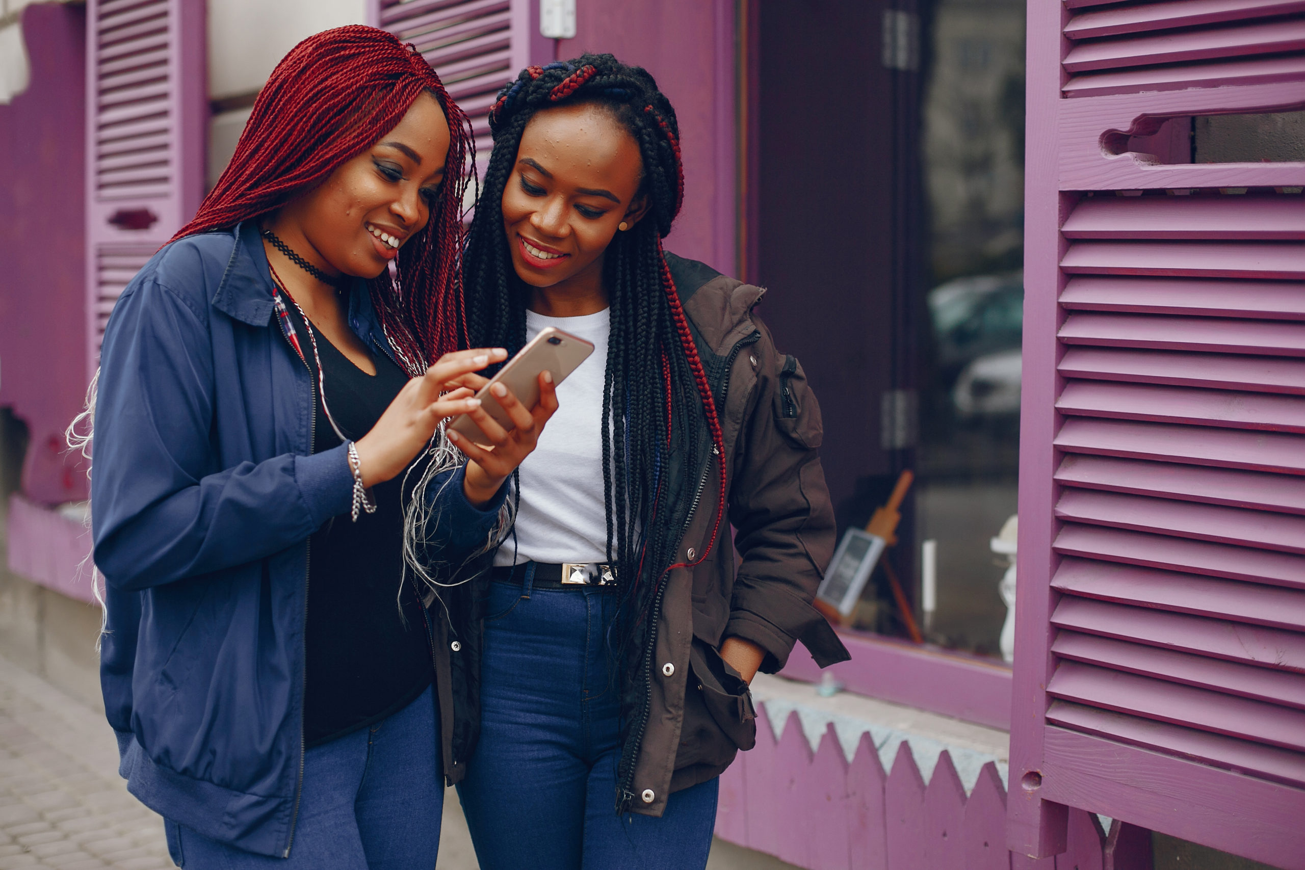 two beautiful and stylish dark-skinned girls with long hair standing in a autumn city near purple wall and using the phone